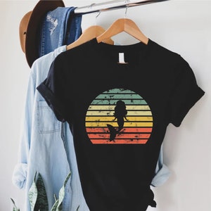 May include: Black t-shirt with a vintage-style sunset graphic featuring a mermaid silhouette. The sunset is in shades of yellow, orange, red, and green.