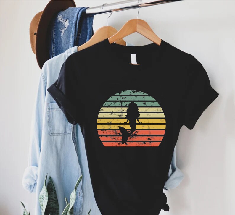 May include: Black t-shirt with a vintage-style sunset graphic featuring a mermaid silhouette. The sunset is in shades of yellow, orange, red, and green.