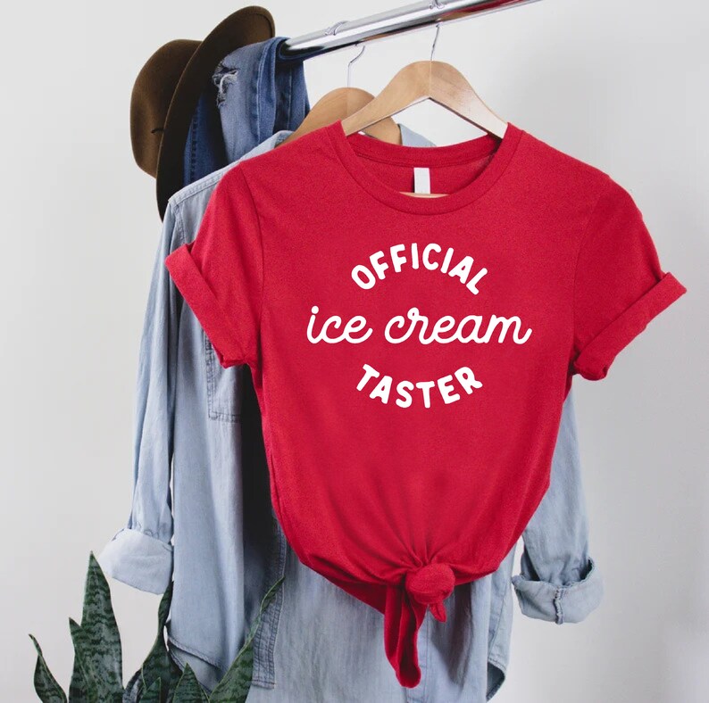 Puede incluir: Camiseta roja con texto blanco que dice "OFFICIAL ice cream TASTER" colgada de una percha.