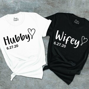 May include: Two white t-shirts with black text. One shirt says "Hubby" with a heart and the date "6.27.20". The other shirt says "Wifey" with a heart and the date "6.27.20".