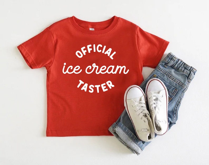 Puede incluir: Camiseta roja de manga corta con texto blanco que dice "OFFICIAL ice cream TASTER". La camiseta se combina con jeans azules y zapatillas blancas.