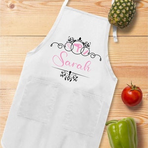 Puede incluir: Delantal blanco con dos bolsillos, con un dise&ntilde;o rosa y negro con el nombre "Sarah". El delantal tiene una correa para el cuello y lazos en la cintura. Una pi&ntilde;a, un tomate y un pimiento verde est&aacute;n sobre la superficie de madera.