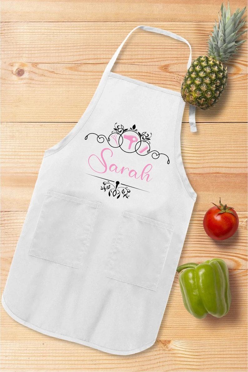 Puede incluir: Delantal blanco con dos bolsillos, con un dise&ntilde;o rosa y negro con el nombre "Sarah". El delantal tiene una correa para el cuello y lazos en la cintura. Una pi&ntilde;a, un tomate y un pimiento verde est&aacute;n sobre la superficie de madera.