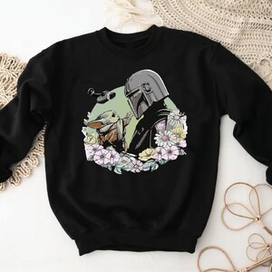 May include: Black sweatshirt featuring a printed design of a Mandalorian character and a Grogu character surrounded by flowers.
