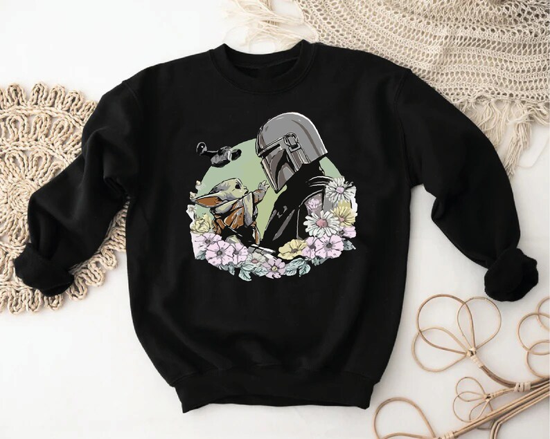 May include: Black sweatshirt featuring a printed design of a Mandalorian character and a Grogu character surrounded by flowers.