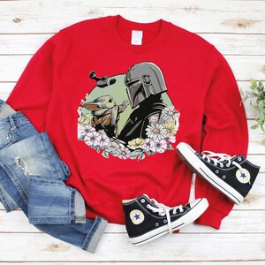 May include: Red sweatshirt featuring a graphic print of a Mandalorian character and a Grogu character surrounded by flowers. The sweatshirt is displayed on a white wooden surface with a pair of blue jeans and black trainers.