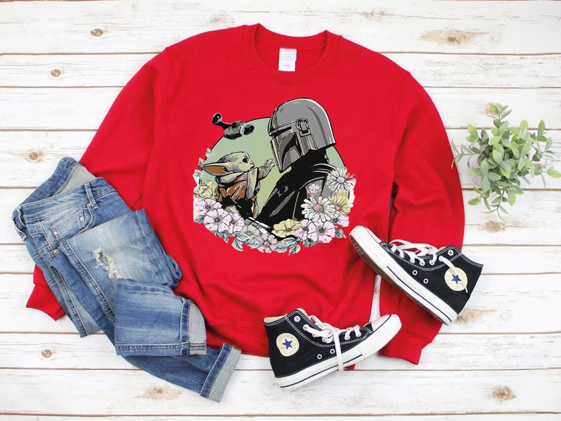 May include: Red sweatshirt featuring a graphic print of a Mandalorian character and a Grogu character surrounded by flowers. The sweatshirt is displayed on a white wooden surface with a pair of blue jeans and black trainers.