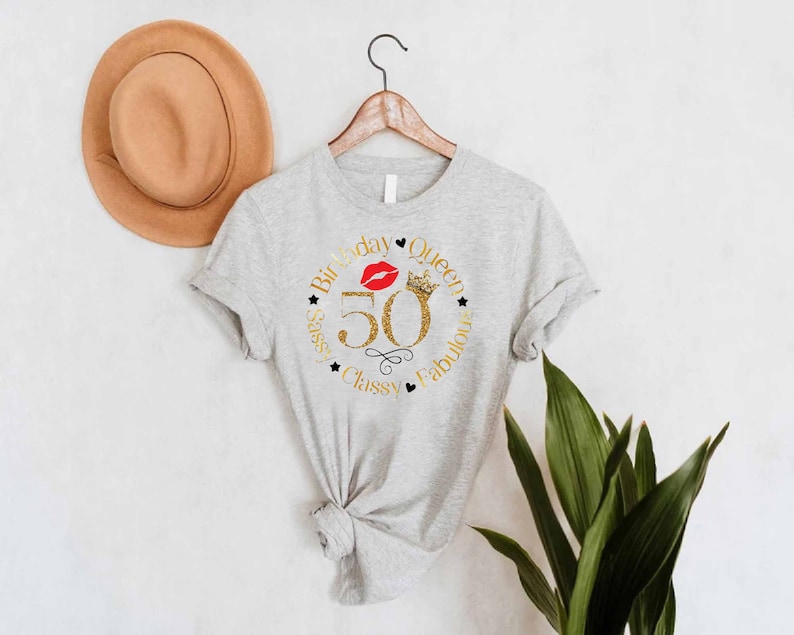 May include: A gray t-shirt with a gold glitter design that says "Birthday Queen" with a red lip and crown. The number 50 is in the center of the design. The words "Sassy, Classy, Fabulous" are around the outside of the design.
