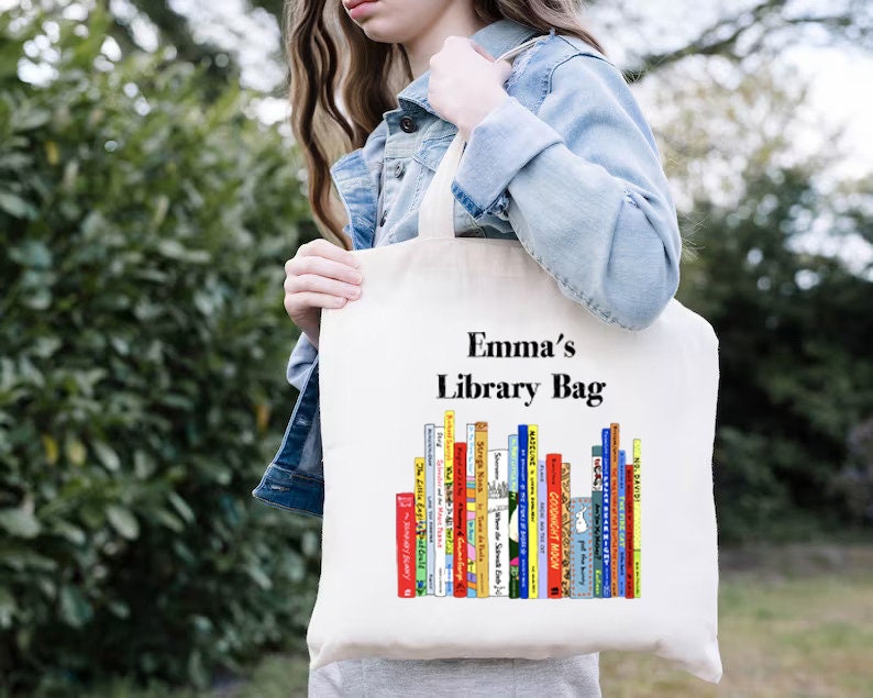 May include: A cream-colored tote bag with the text "Emma's Library Bag" above a colorful illustration of stacked books. The bag is held by a person wearing a denim jacket, with a green leafy background.