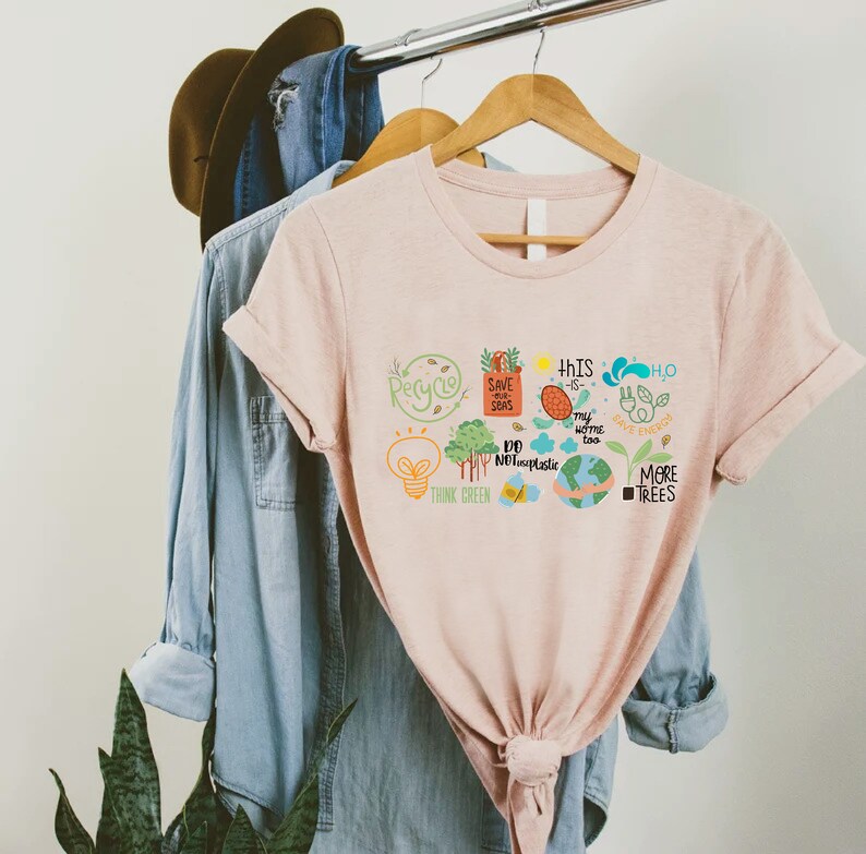 Puede incluir: Una camiseta rosa con un dise&ntilde;o gr&aacute;fico que presenta una bombilla, &aacute;rboles, un globo terr&aacute;queo y texto que dice "Recicla", "Salva nuestros mares", "No al pl&aacute;stico", "Piensa en verde", "Este tambi&eacute;n es mi hogar", "H2O", "Ahorra energ&iacute;a" y "M&aacute;s &aacute;rboles".
