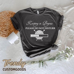 May include: A dark gray t-shirt with white text that reads "Knitting in Progress Please Do Not Disturb" and a graphic of knitting needles and yarn with a progress bar.