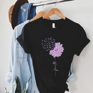 May include: Black t-shirt with a purple flower design. The flower has purple ribbons blowing away from it. The word "faith" is written vertically below the flower.