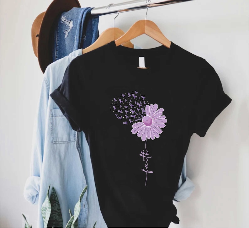 May include: Black t-shirt with a purple flower design. The flower has purple ribbons blowing away from it. The word "faith" is written vertically below the flower.