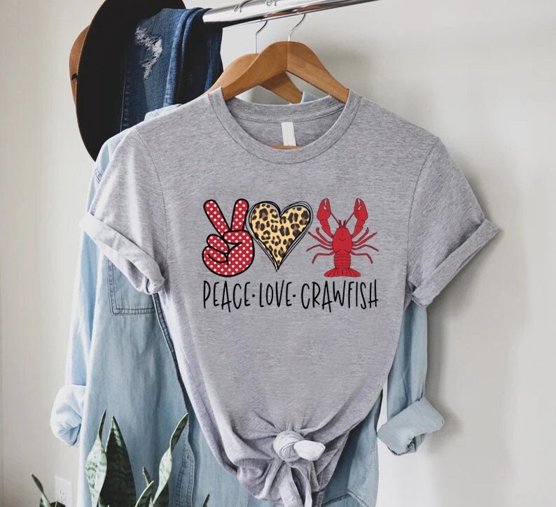 May include: Gray t-shirt with a peace sign, heart, and crawfish graphic. The heart is leopard print and the peace sign has red polka dots. The text reads "Peace-Love-Crawfish".