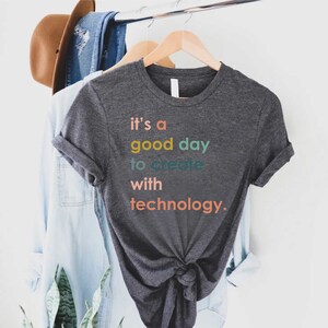 May include: A dark grey t-shirt with the text "it's a good day to create with technology." printed in colourful letters.