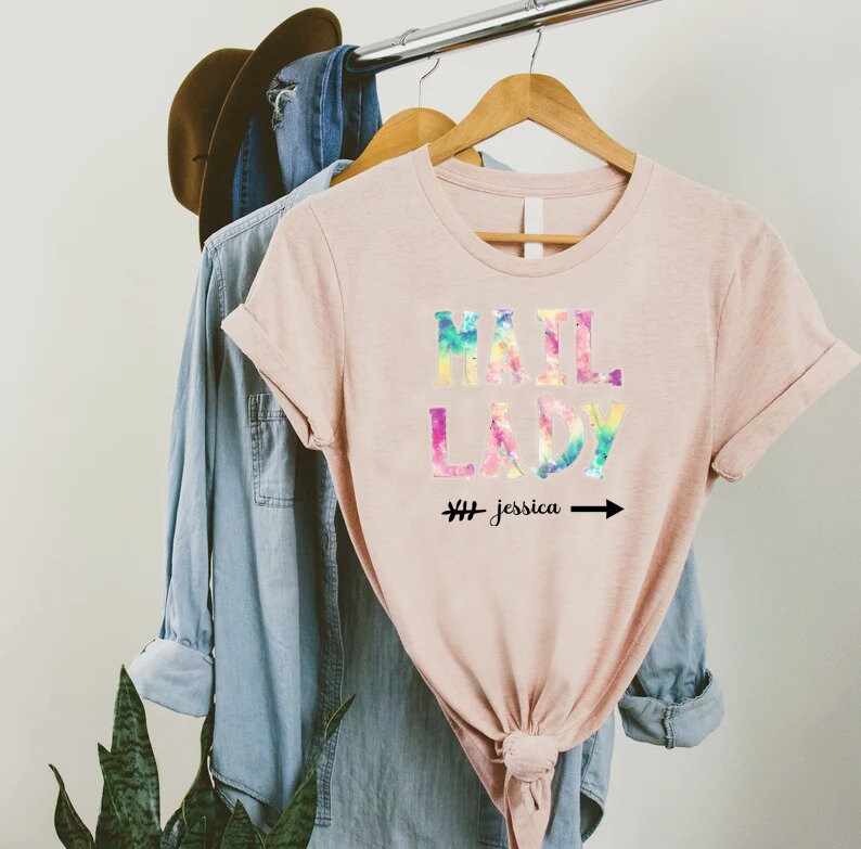 May include: A pink t-shirt with the words "Mail Lady" in rainbow tie-dye lettering. The text "# Jessica" and an arrow are below the words.