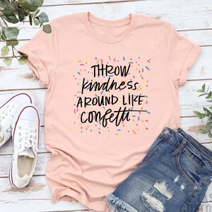 Może przedstawiać: R&oacute;żowy T-shirt z napisem "Throw kindness around like confetti" czarnymi literami. Tekst jest otoczony kolorowymi konfetti.