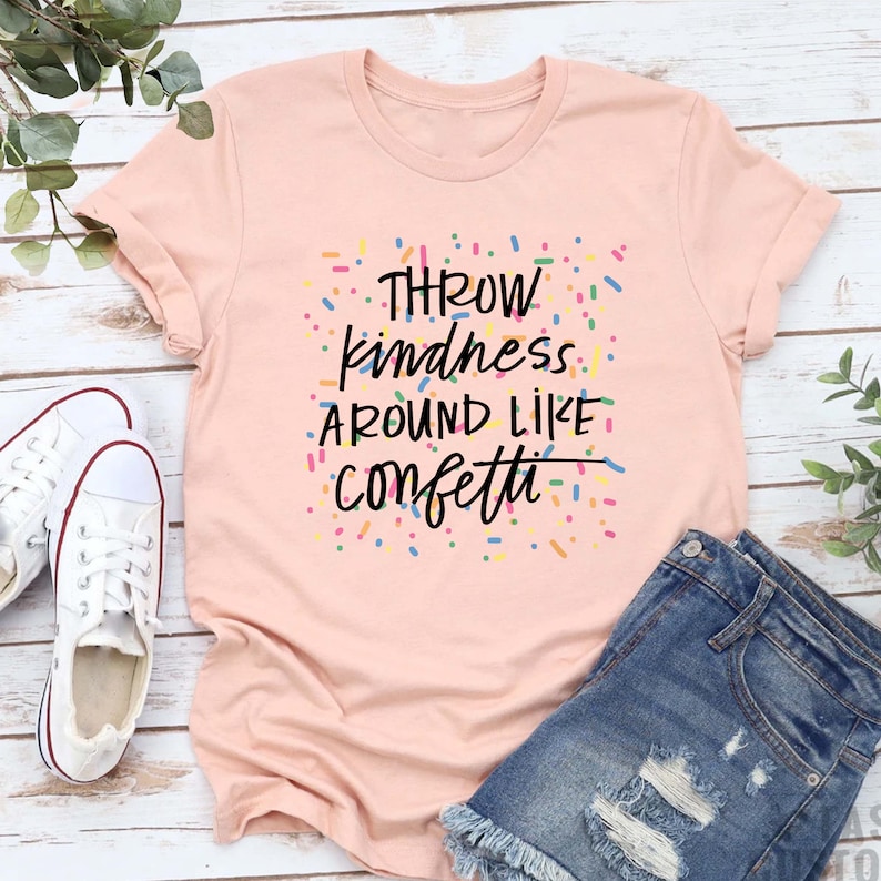 Może przedstawiać: R&oacute;żowy T-shirt z napisem "Throw kindness around like confetti" czarnymi literami. Tekst jest otoczony kolorowymi konfetti.
