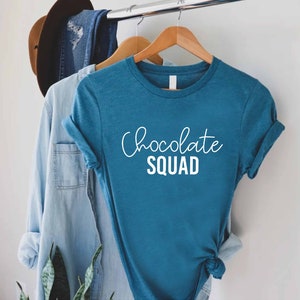 Peut inclure: Un t-shirt bleu sarcelle avec le texte "Chocolate Squad" en lettres blanches.