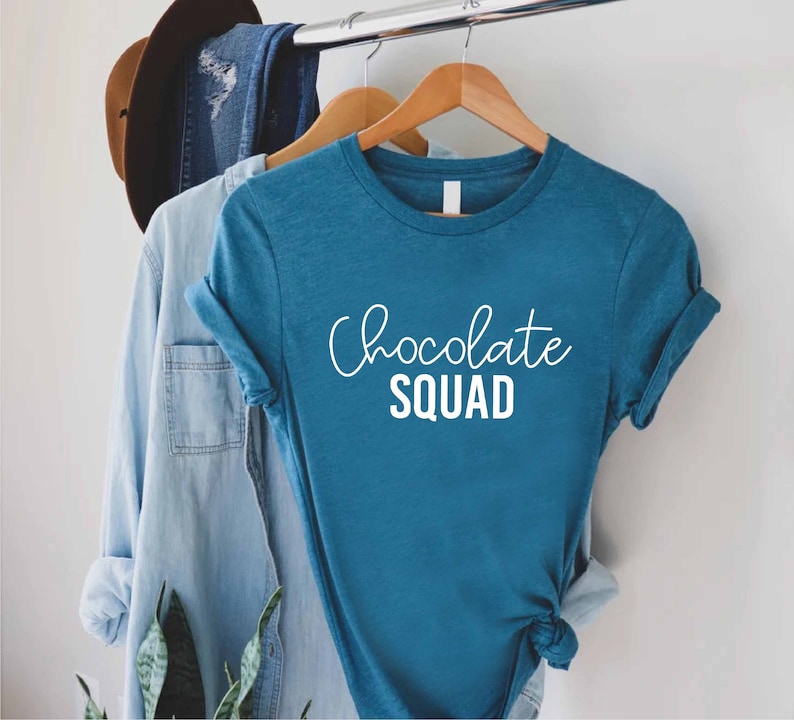 Peut inclure: Un t-shirt bleu sarcelle avec le texte "Chocolate Squad" en lettres blanches.