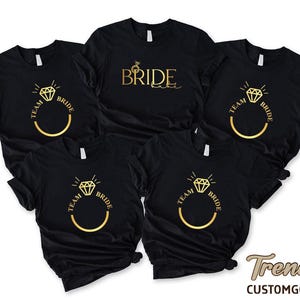 May include: Five black t-shirts with gold text and graphics. One shirt says "BRIDE" with a ring graphic. The other four shirts say "TEAM BRIDE" with a ring graphic. The shirts are short-sleeved.