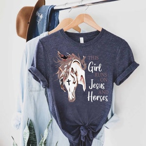 May include: A navy blue t-shirt with a graphic of a horse's head with a cross on its forehead. The text "This Girl Runs On Jesus And Horses" is printed on the shirt.