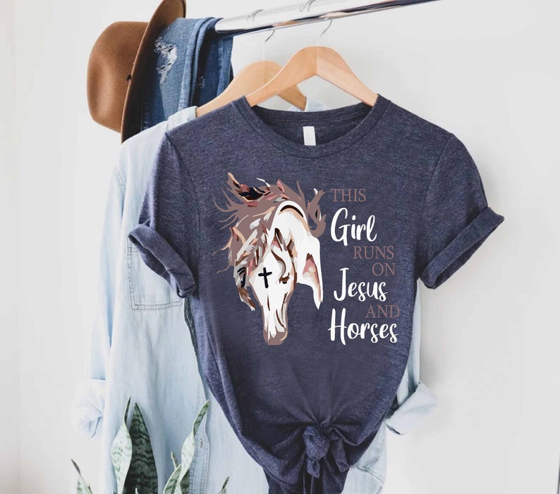 May include: A navy blue t-shirt with a graphic of a horse's head with a cross on its forehead. The text "This Girl Runs On Jesus And Horses" is printed on the shirt.