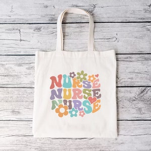 Puede incluir: Una bolsa de lona blanca con un dise&ntilde;o retro colorido que dice "NURSE" rodeada de flores y corazones.