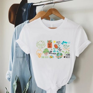 Puede incluir: Camiseta blanca con un dise&ntilde;o gr&aacute;fico que presenta una variedad de iconos y textos ecol&oacute;gicos, incluyendo "Reciclar", "Salvar nuestros mares", "Piensa en verde", "Este tambi&eacute;n es mi hogar", "No uses pl&aacute;stico", "H2O Ahorra energ&iacute;a", y "M&aacute;s &aacute;rboles".