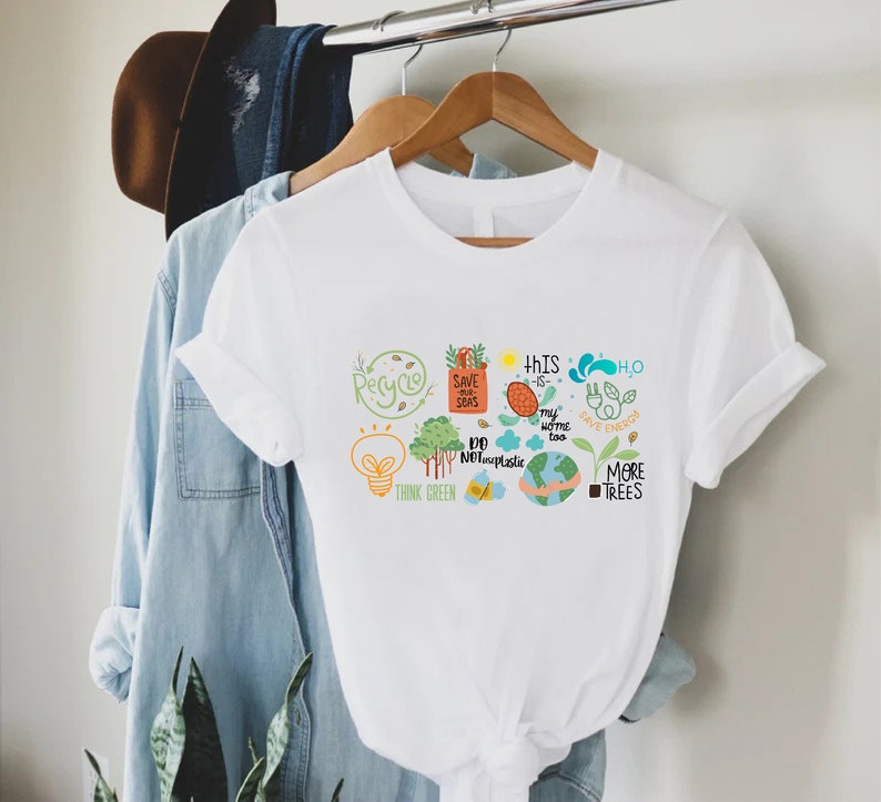 Puede incluir: Camiseta blanca con un dise&ntilde;o gr&aacute;fico que presenta una variedad de iconos y textos ecol&oacute;gicos, incluyendo "Reciclar", "Salvar nuestros mares", "Piensa en verde", "Este tambi&eacute;n es mi hogar", "No uses pl&aacute;stico", "H2O Ahorra energ&iacute;a", y "M&aacute;s &aacute;rboles".