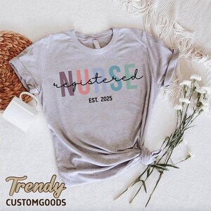 K&ouml;nnte beinhalten: Ein graues T-Shirt mit dem Text "NURSE registered EST. 2025" in Regenbogenfarben. Der Text ist in einer kursivschrift.