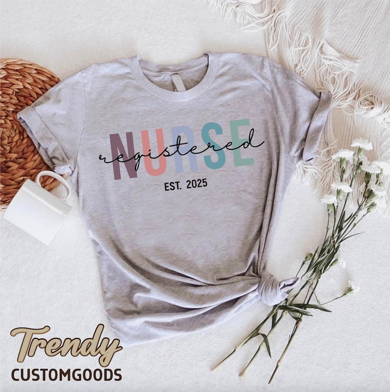 K&ouml;nnte beinhalten: Ein graues T-Shirt mit dem Text "NURSE registered EST. 2025" in Regenbogenfarben. Der Text ist in einer kursivschrift.