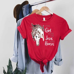 May include: Red t-shirt with a white horse head graphic and the text "This Girl Runs On Jesus And Horses".