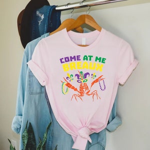 Może przedstawiać: R&oacute;żowa koszulka z rakiem w masce Mardi Gras i napisem "Come at me Breaux".