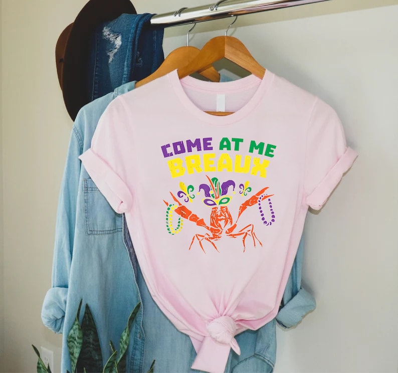 Może przedstawiać: R&oacute;żowa koszulka z rakiem w masce Mardi Gras i napisem "Come at me Breaux".