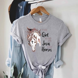 May include: Gray t-shirt with a graphic of a horse's head with a cross on its forehead. The text "This Girl Runs On Jesus And Horses" is printed on the shirt.