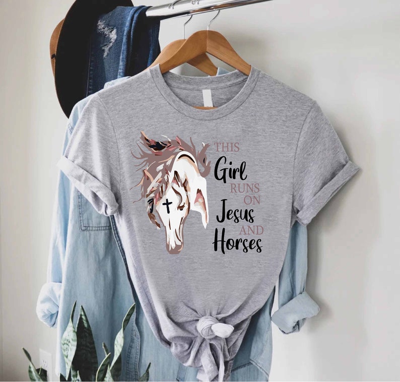 May include: Gray t-shirt with a graphic of a horse's head with a cross on its forehead. The text "This Girl Runs On Jesus And Horses" is printed on the shirt.