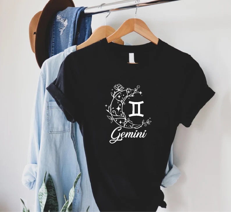Puede incluir: Camiseta negra con un gr&aacute;fico blanco de una luna creciente con estrellas y enredaderas florales que rodean el s&iacute;mbolo del zod&iacute;aco G&eacute;minis. El texto "G&eacute;minis" est&aacute; escrito debajo del s&iacute;mbolo.