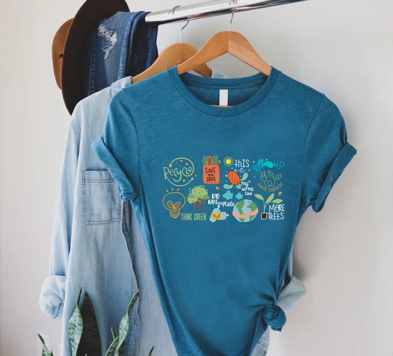 Puede incluir: Una camiseta azul turquesa con un estampado gr&aacute;fico de varios lemas e ilustraciones ambientales, incluyendo "Recicla", "Salva nuestros mares", "Piensa en verde", "No uses pl&aacute;stico", "M&aacute;s &aacute;rboles" y "Ahorra energ&iacute;a".
