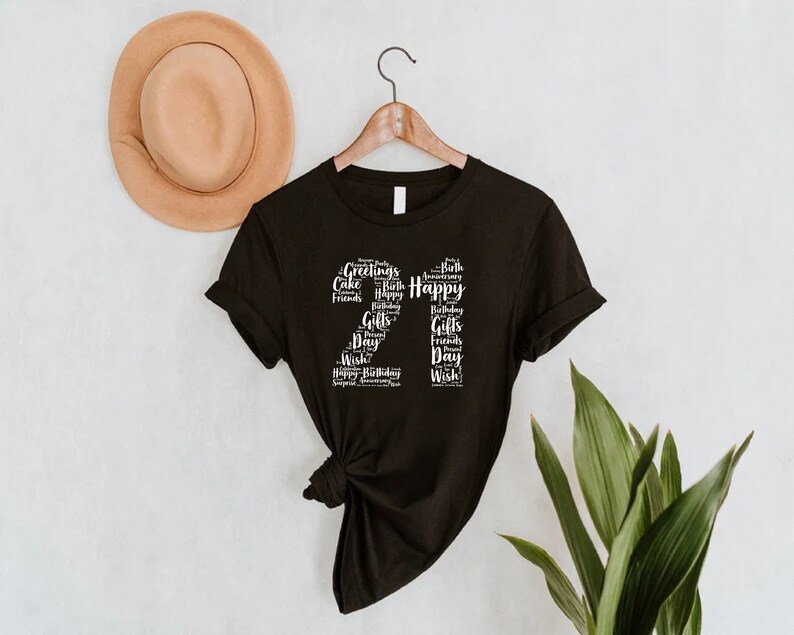 May include: Black t-shirt with a white graphic of the number 21 made up of words related to birthdays, such as "Happy", "Birthday", "Cake", "Friends", and "Gifts".