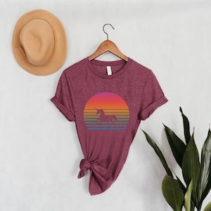 K&ouml;nnte beinhalten: Burgunderfarbenes T-Shirt mit einem farbenfrohen Sonnenuntergangsdesign mit einer Einhorn-Silhouette. Die Grafik enth&auml;lt einen Farbverlauf aus Orange, Rosa und Gelb mit blauen und gr&uuml;nen Streifen darunter. Das Shirt h&auml;ngt an einem Holzb&uuml;gel.