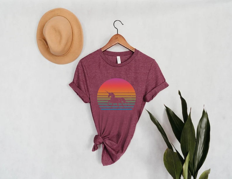 K&ouml;nnte beinhalten: Burgunderfarbenes T-Shirt mit einem farbenfrohen Sonnenuntergangsdesign mit einer Einhorn-Silhouette. Die Grafik enth&auml;lt einen Farbverlauf aus Orange, Rosa und Gelb mit blauen und gr&uuml;nen Streifen darunter. Das Shirt h&auml;ngt an einem Holzb&uuml;gel.
