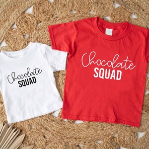 Peut inclure: Deux t-shirts avec le texte "Chocolate Squad" imprim&eacute; dessus. Un t-shirt est blanc avec du texte noir et l'autre est rouge avec du texte blanc.
