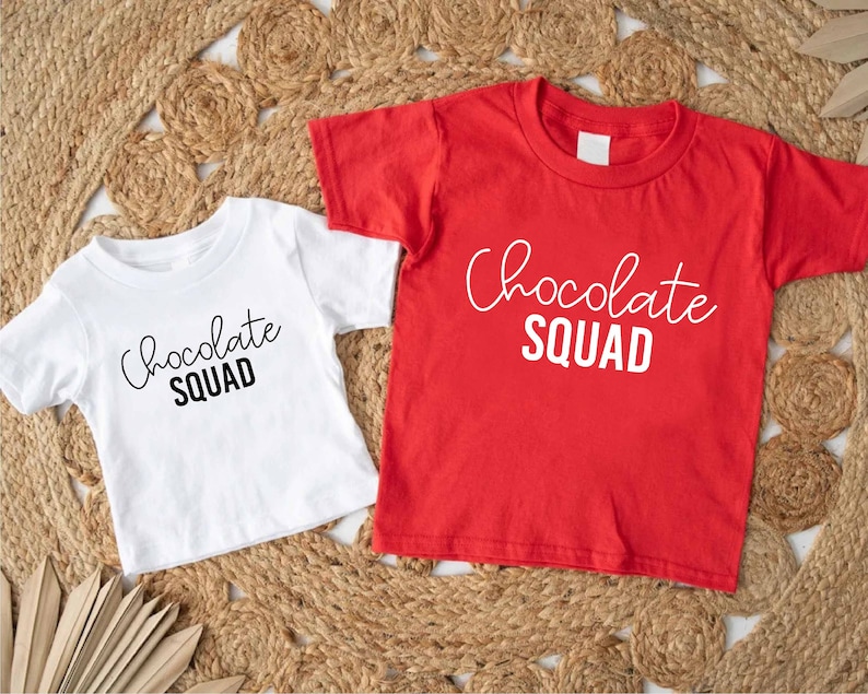 Peut inclure: Deux t-shirts avec le texte "Chocolate Squad" imprim&eacute; dessus. Un t-shirt est blanc avec du texte noir et l'autre est rouge avec du texte blanc.