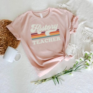May include: A pink t-shirt with the text "History Teacher" in a retro font with a rainbow stripe design.