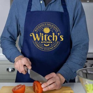 May include: A blue apron with a yellow circle design that says "Wickedly Delicious Witch's Kitchen Stirring Up Some Magic". A person is wearing the apron and holding a knife.