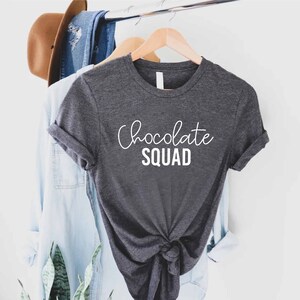Peut inclure: Un t-shirt gris fonc&eacute; avec le texte "Chocolate Squad" en lettres blanches.