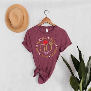 May include: A maroon t-shirt with a gold glitter design that says "Birthday Queen Sassy, Classy, Fabulous" with a red lip and crown inside the circle. The number 50 is in the center of the circle.