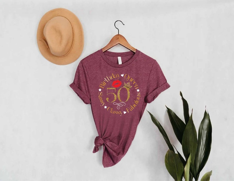 May include: A maroon t-shirt with a gold glitter design that says "Birthday Queen Sassy, Classy, Fabulous" with a red lip and crown inside the circle. The number 50 is in the center of the circle.