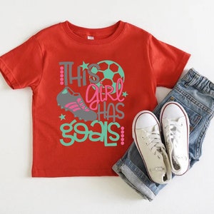 May include: Red short-sleeved t-shirt with the text "This girl has goals" in green and pink lettering. A soccer ball and a soccer cleat are also featured on the shirt.